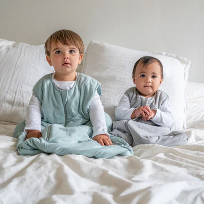 Two children sitting on a bed wearing light-colored clothing.