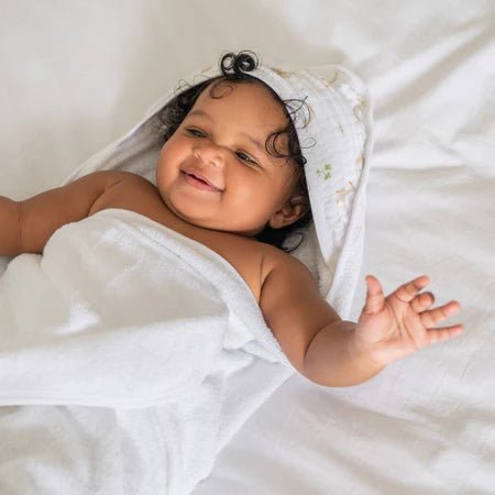 Baby wrapped in Aden + Anais Essentials Dino Jungle hooded cotton muslin towel on white blanket