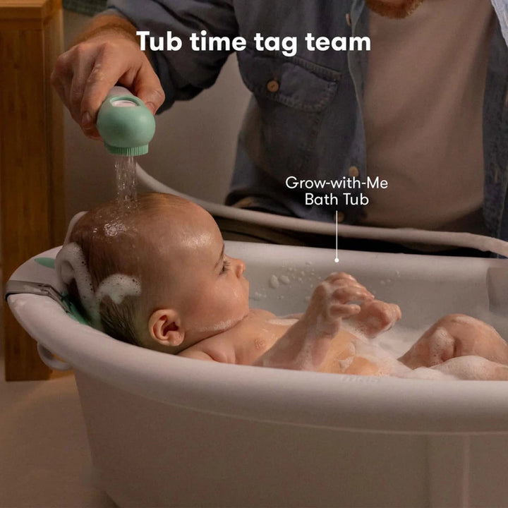 Fridababy - Control the Flow Bath Sprayer -  A baby sits in a white bath tub while an adult showers the baby  