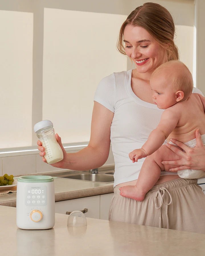 Momcozy Nutri Smart Analog Baby Bottle Warmer on kitchen counter with mother feeding baby using warmed milk bottle
