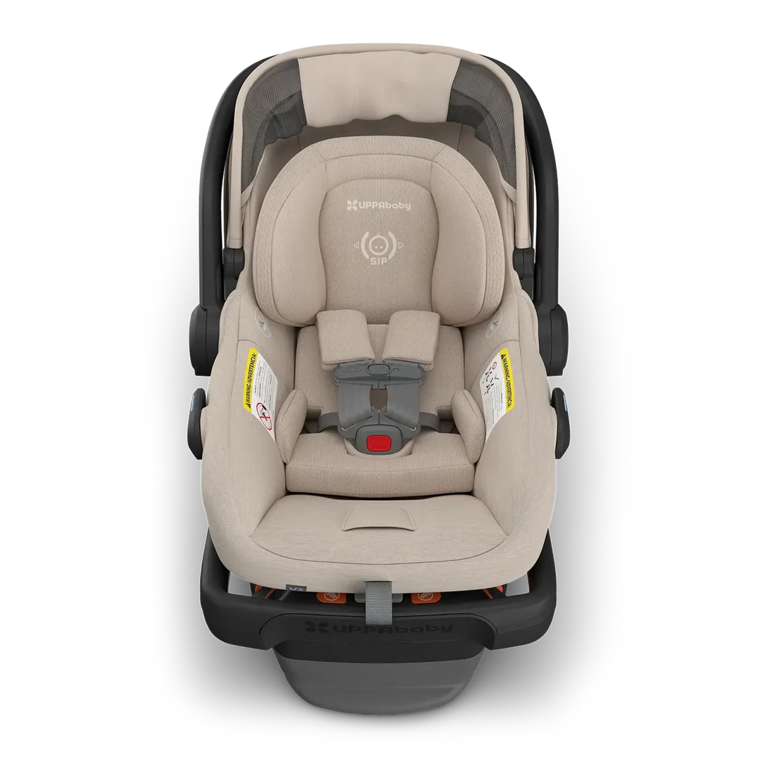 A UPPAbaby infant car seat in a light beige fabric with a dark gray and black base, shown from a top-down view on a white background.