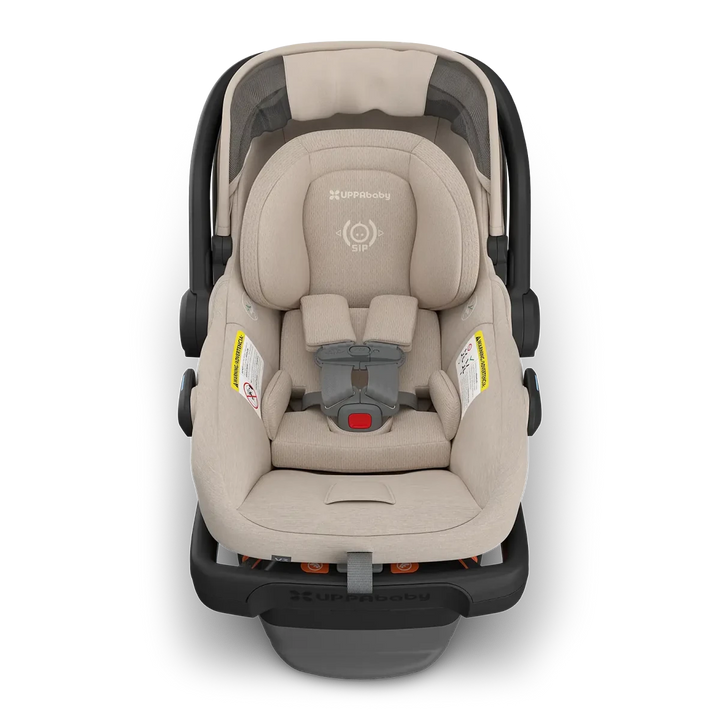 A UPPAbaby infant car seat in a light beige fabric with a dark gray and black base, shown from a top-down view on a white background.